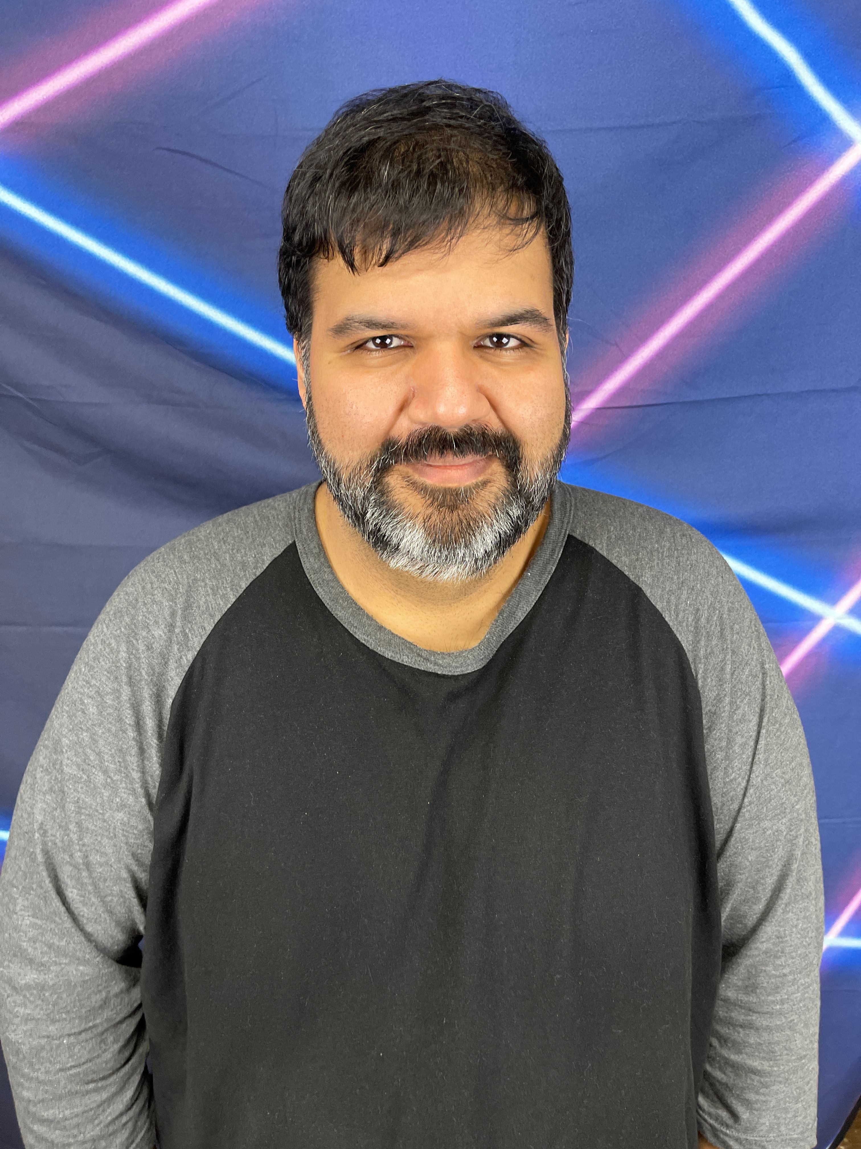 Man with dark hair and beard wearing a black and gray long-sleeve shirt, smiling, with colorful neon lights in the background.