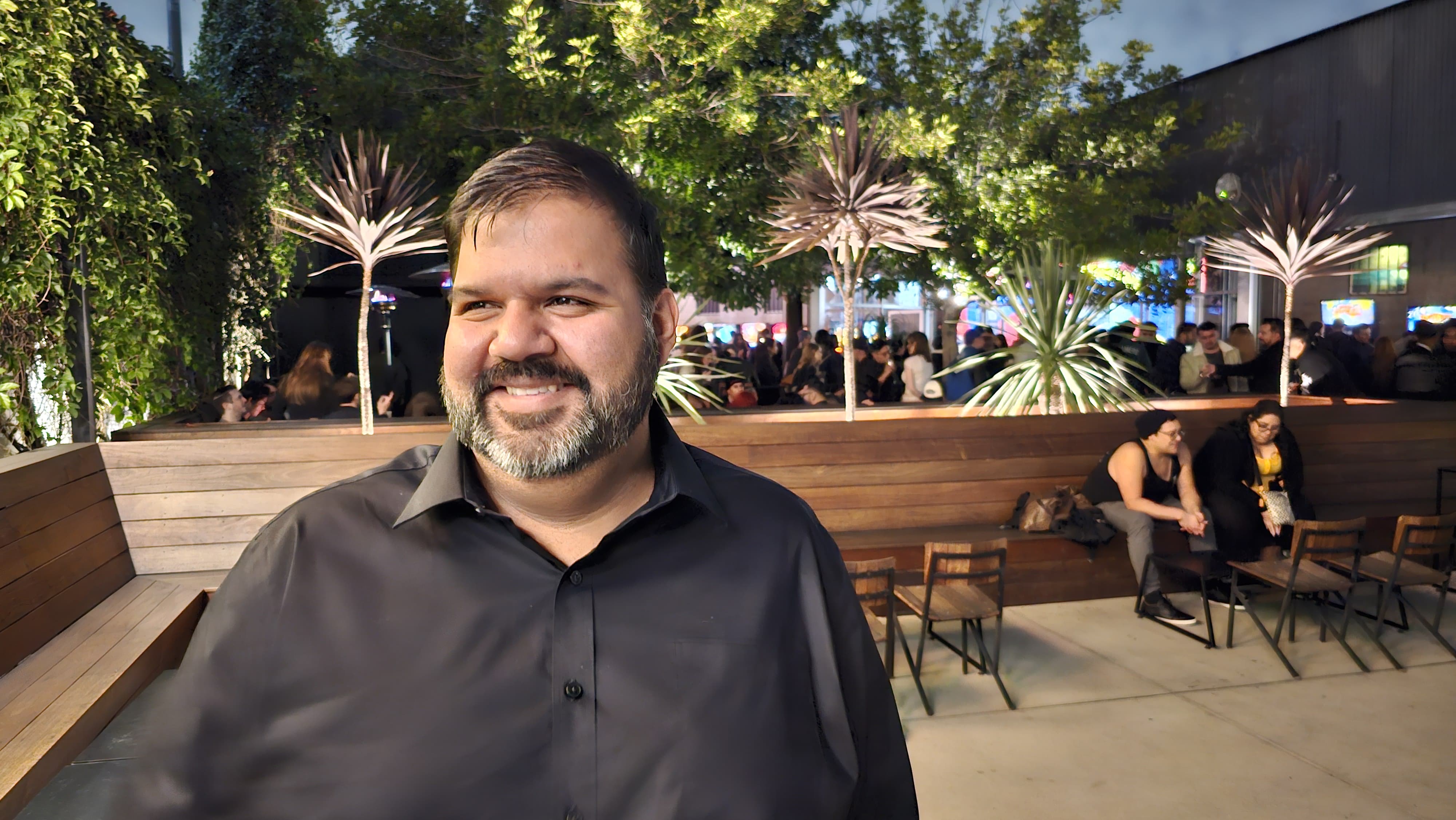 Man with beard smiling outdoors at night, trees and people in background, seated at tables.