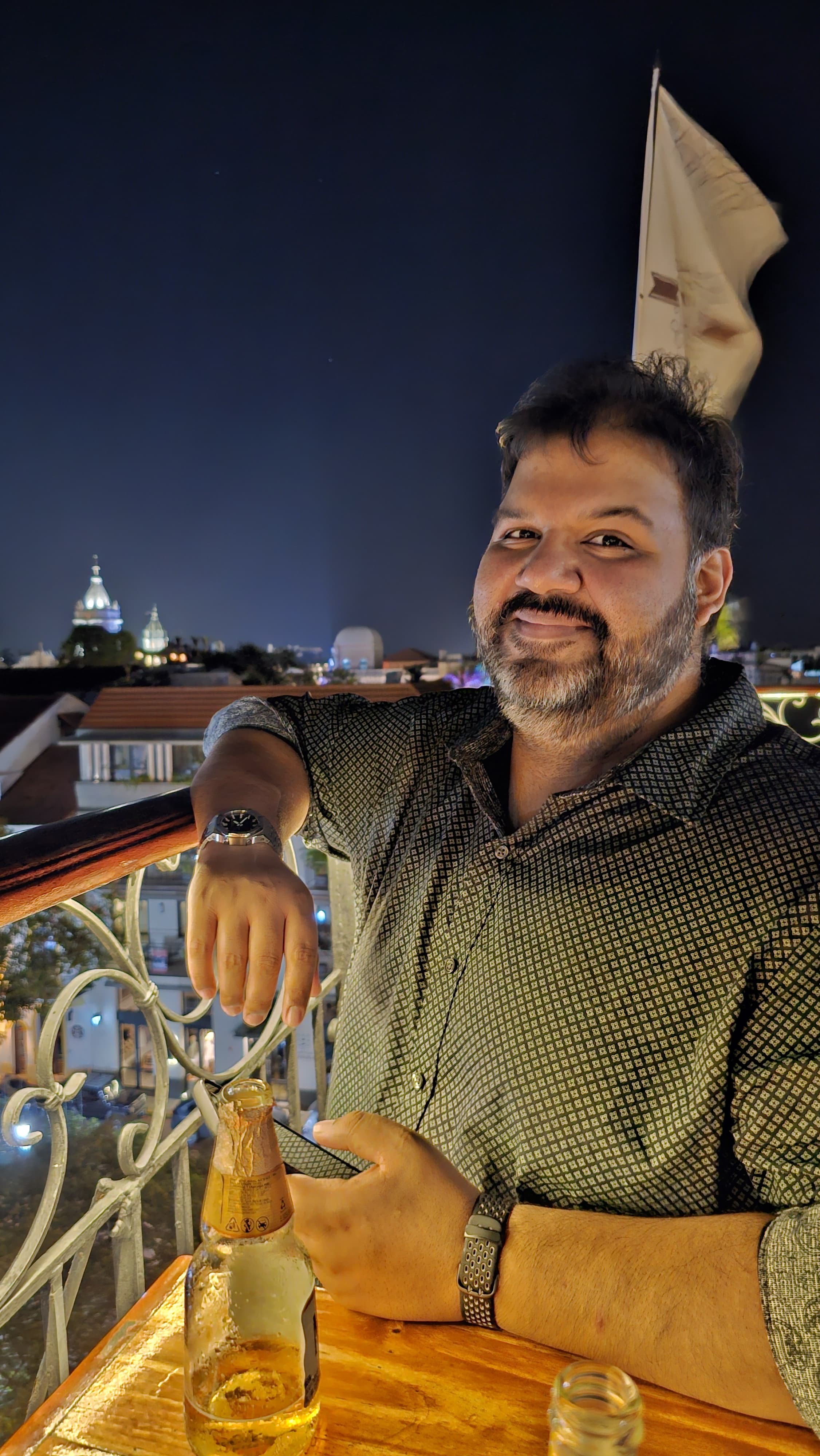Man with beard and short hair smiling, sitting at a table with a bottle, night cityscape background with illuminated buildings.