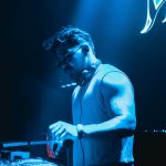 DJ wearing sunglasses and a sleeveless shirt, operating equipment in a blue-lit environment.