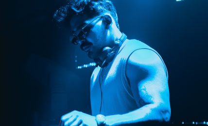 DJ wearing sunglasses and a sleeveless shirt, operating equipment in a blue-lit environment.