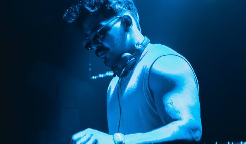 DJ wearing sunglasses and a sleeveless shirt, operating equipment in a blue-lit environment.