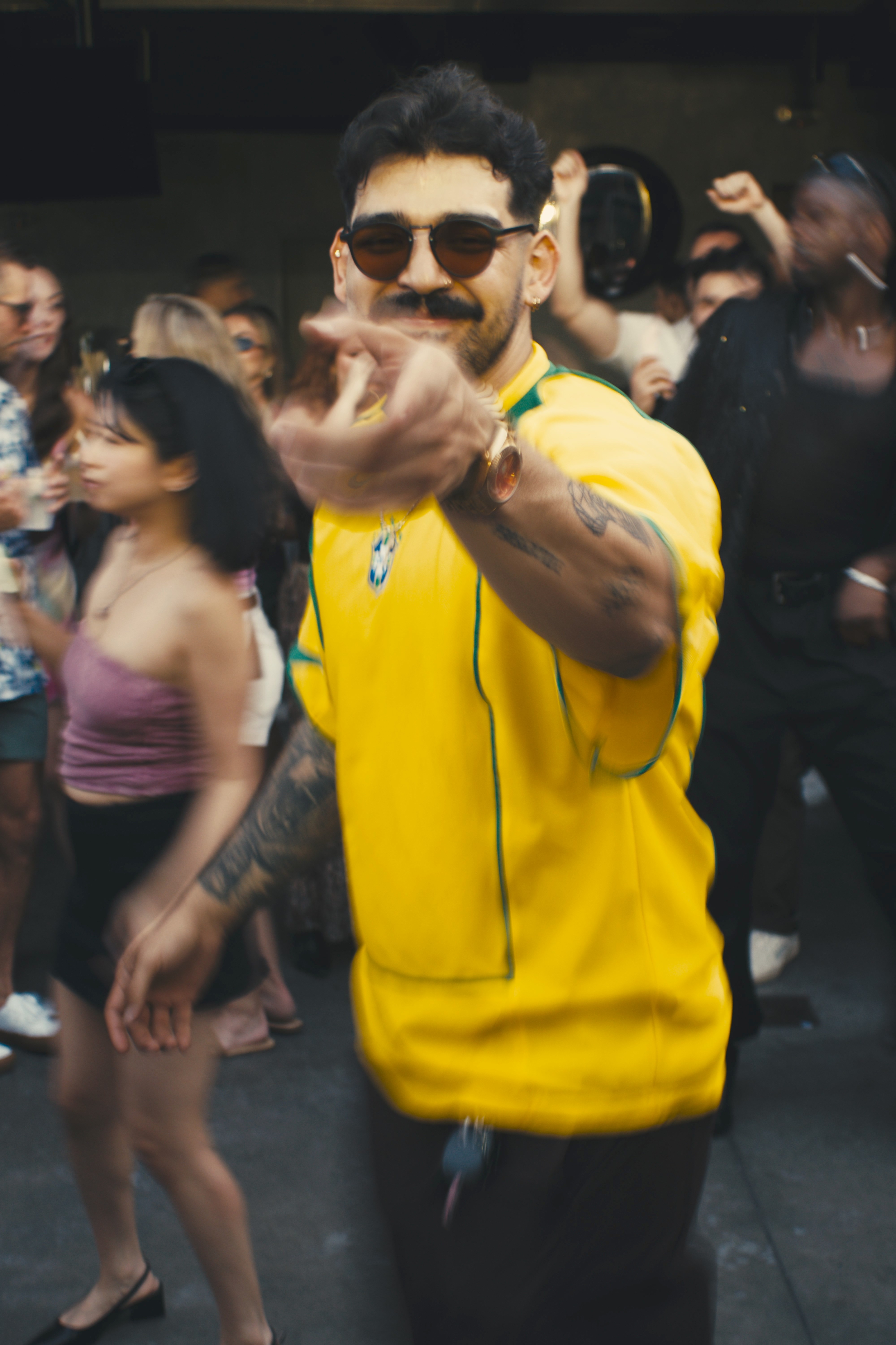 Man with sunglasses smiling and pointing at camera, surrounded by people in a social setting.