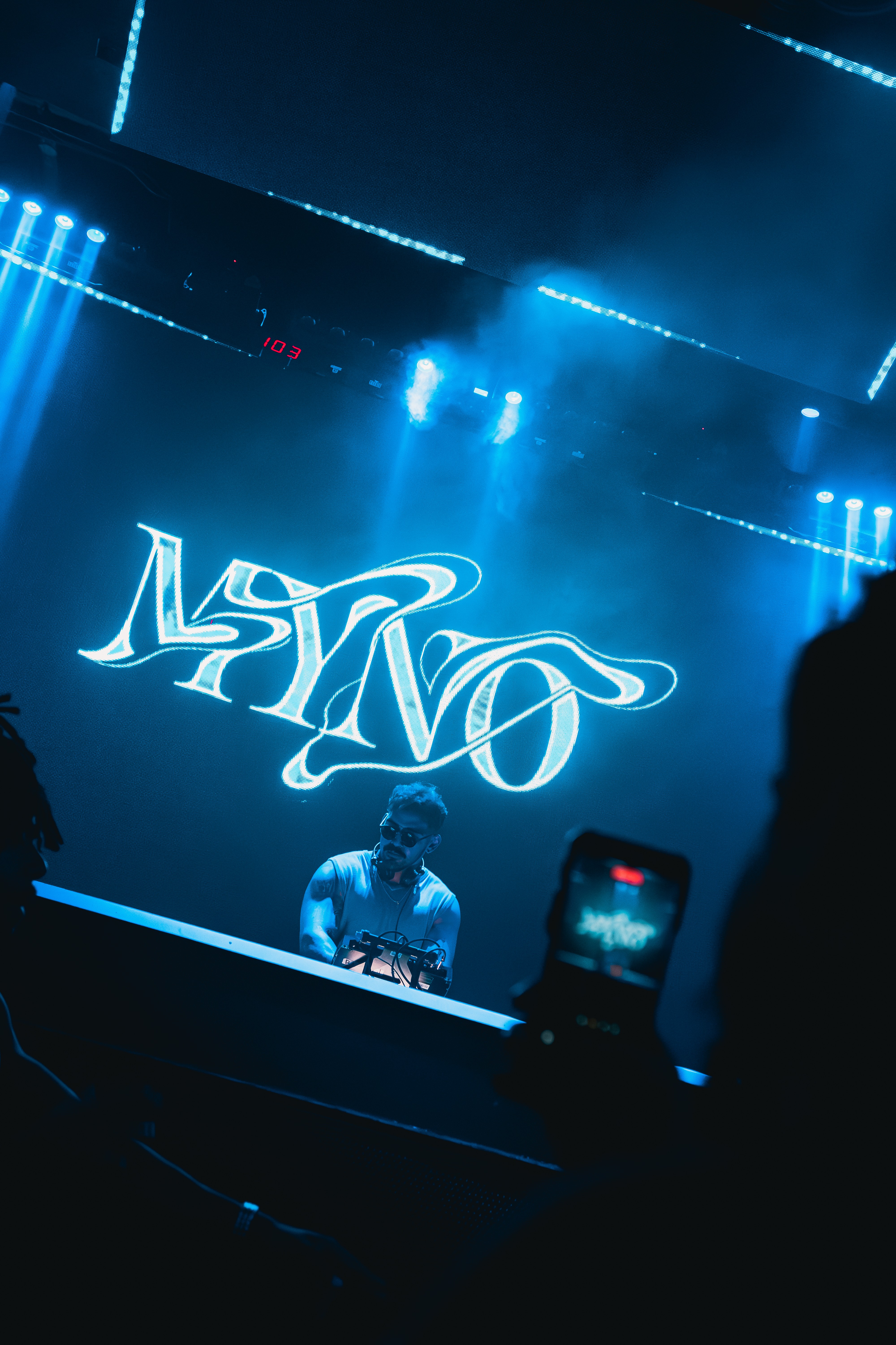 A DJ performs on stage with blue lighting and a large illuminated logo behind him, audience members visible in foreground.