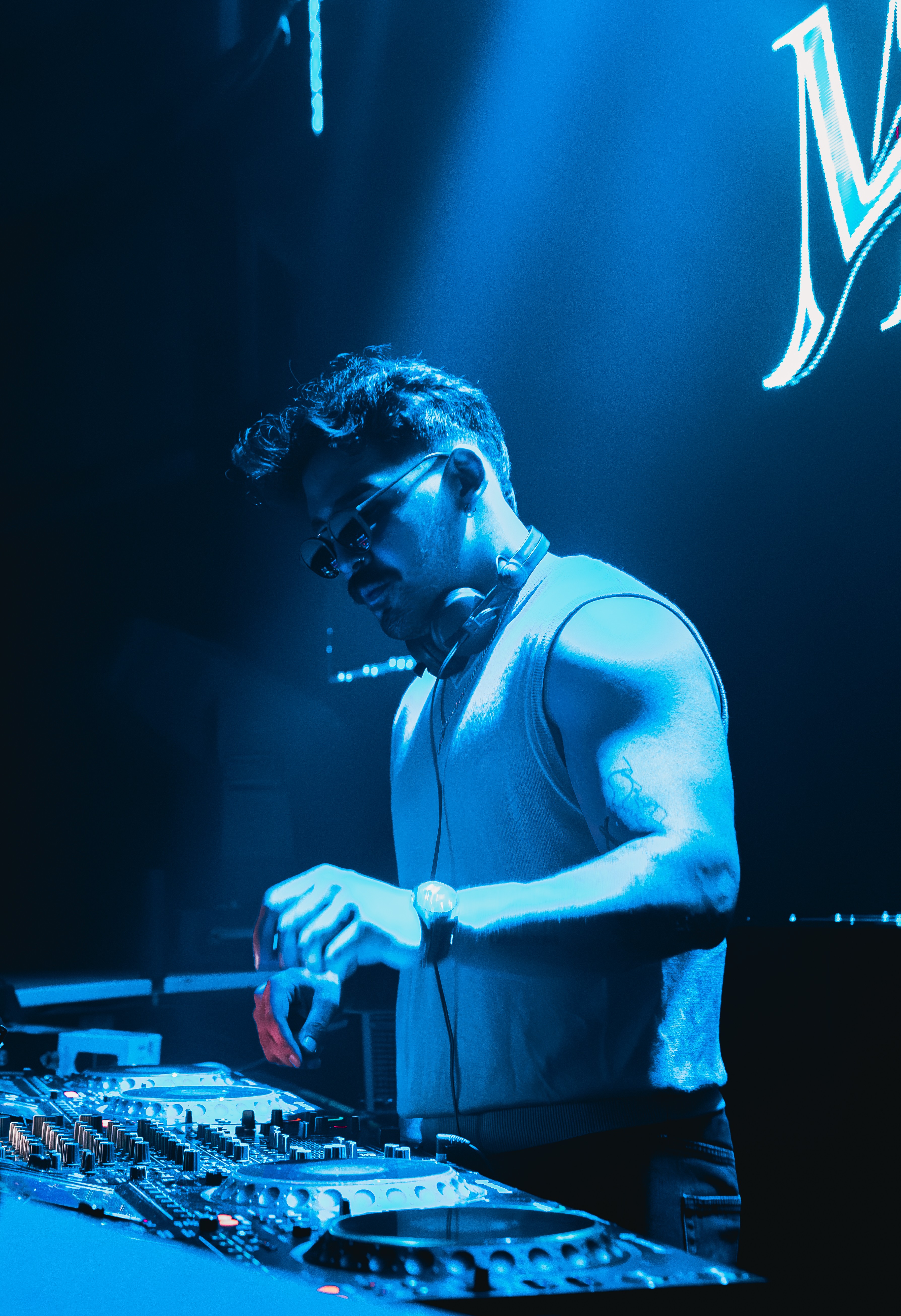 DJ wearing sunglasses and sleeveless shirt, operating equipment in a blue-lit environment, with a large illuminated sign in the background.