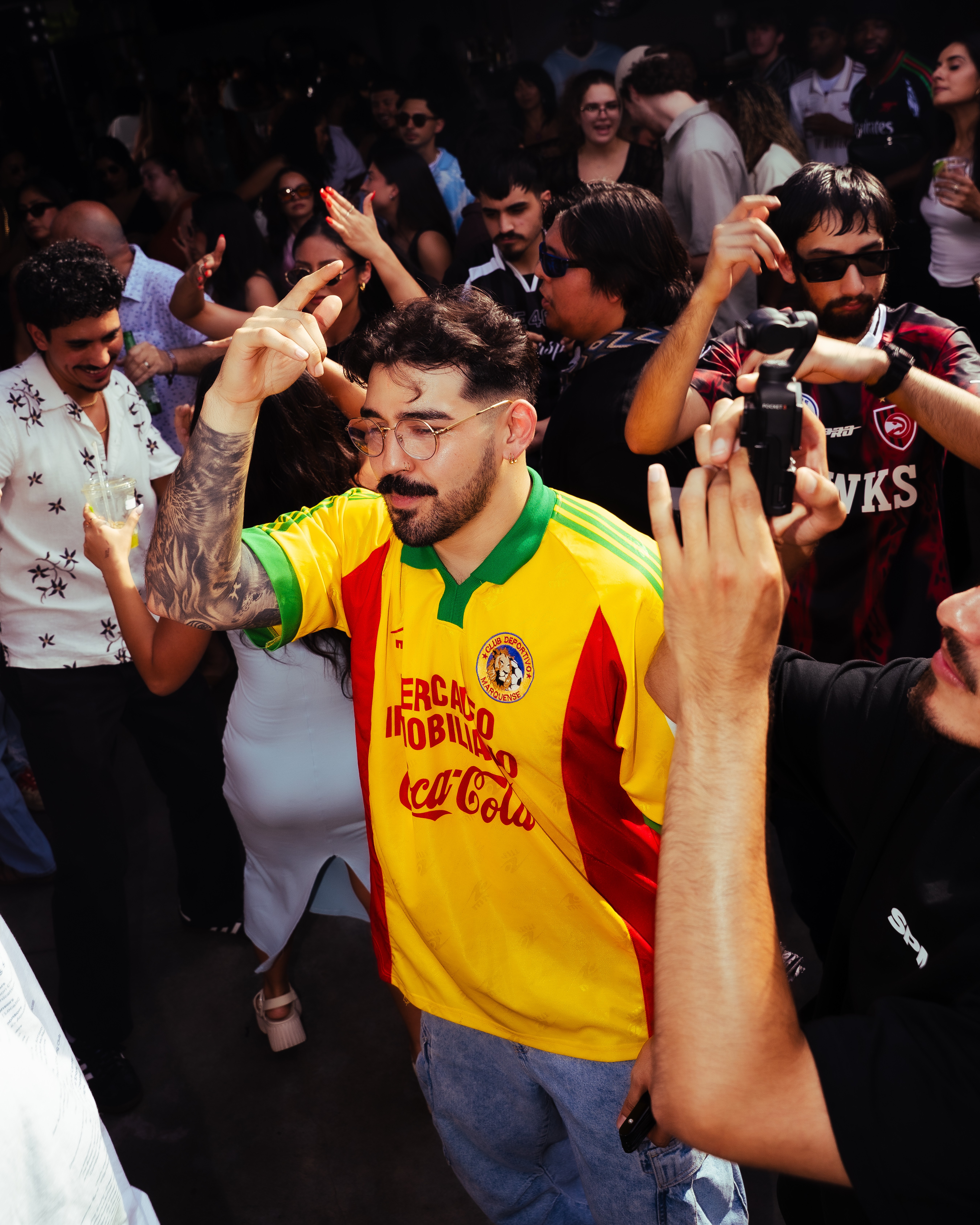 Man with glasses and tattoos wearing a yellow and green sports jersey, raising his hand in a crowd.