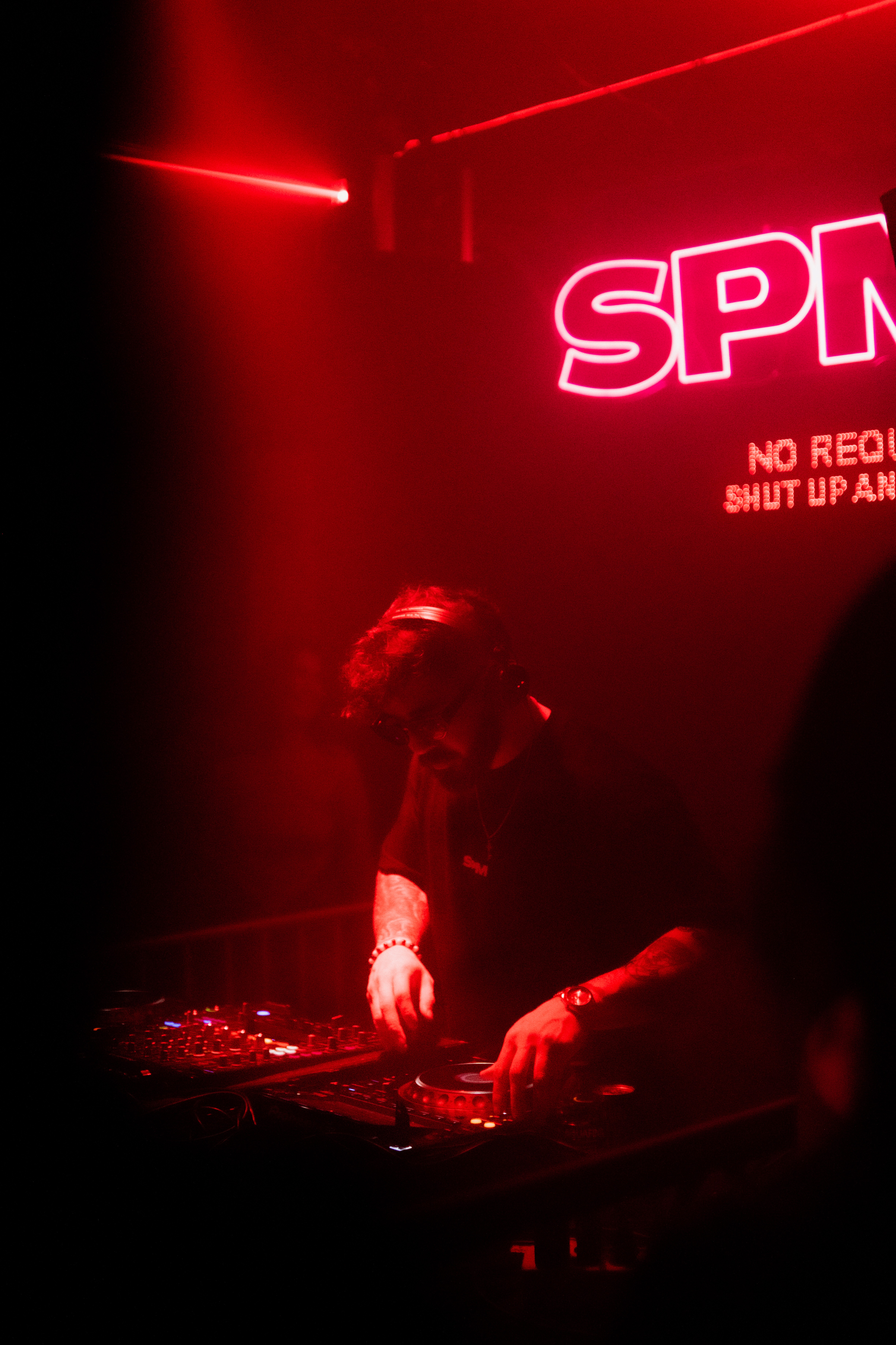 Person DJing in a dark, red-lit club with a neon sign reading 'SPN'.
