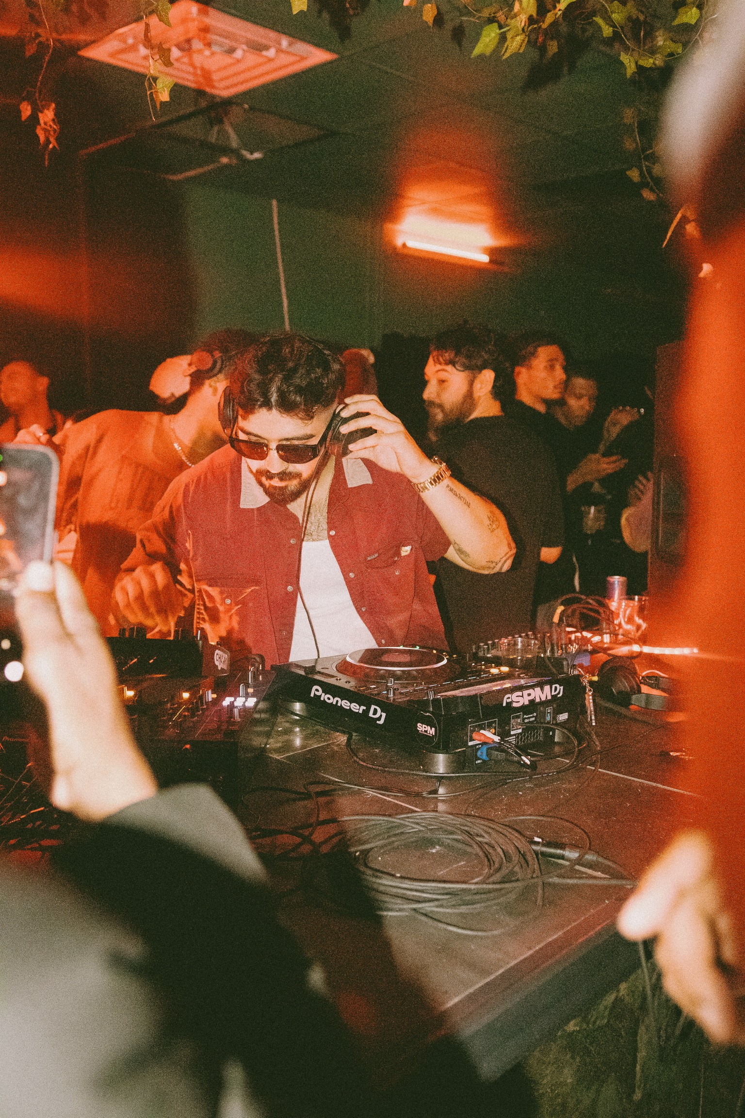 DJ with sunglasses and headphones performing at a crowded party with colorful lighting.