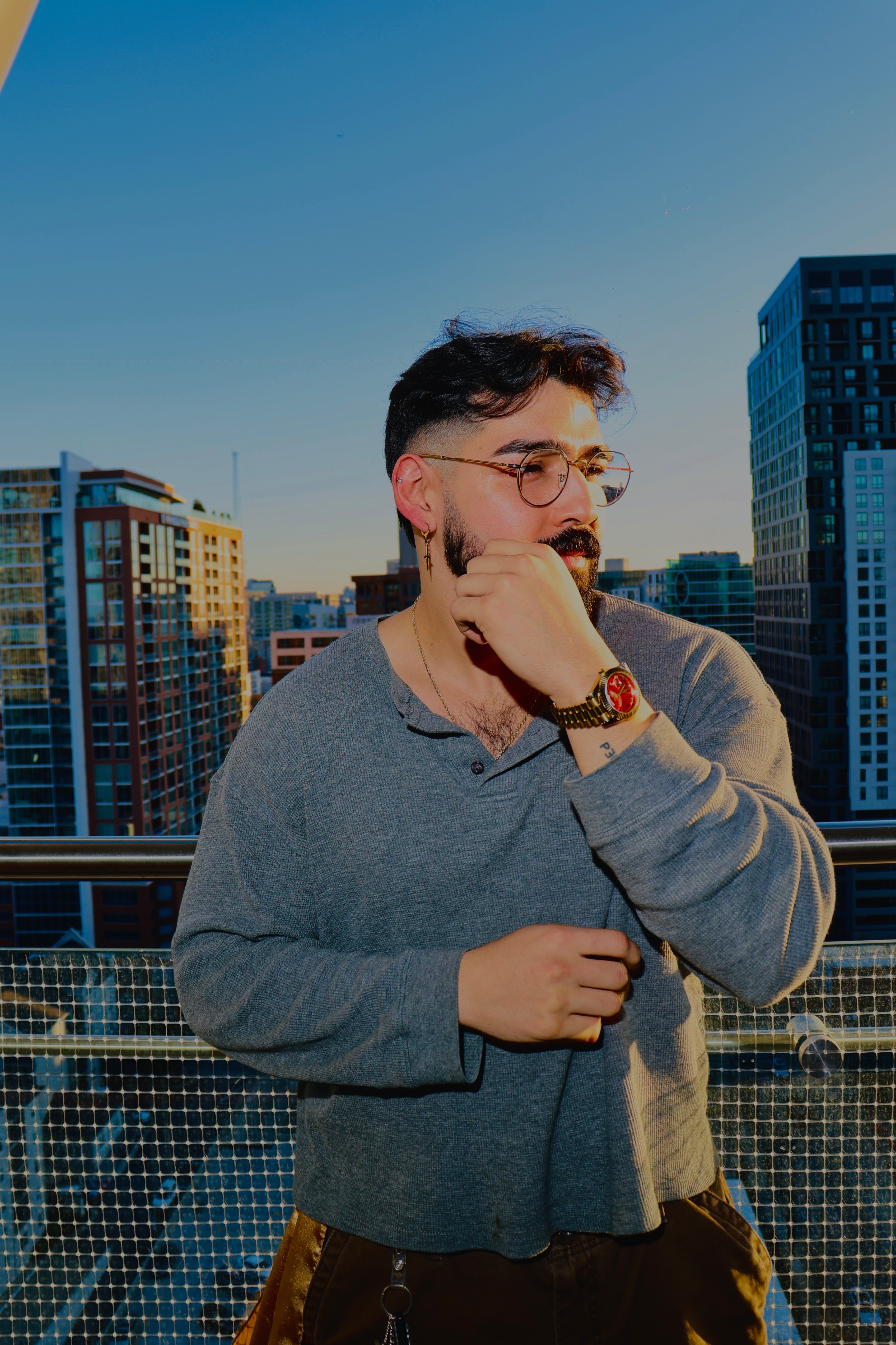 Man with glasses and a beard wearing a gray sweater, standing outdoors in a cityscape with tall buildings, during daytime.