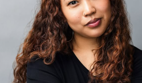Portrait of a woman with long, curly hair wearing a black top, looking at the camera with a neutral expression.