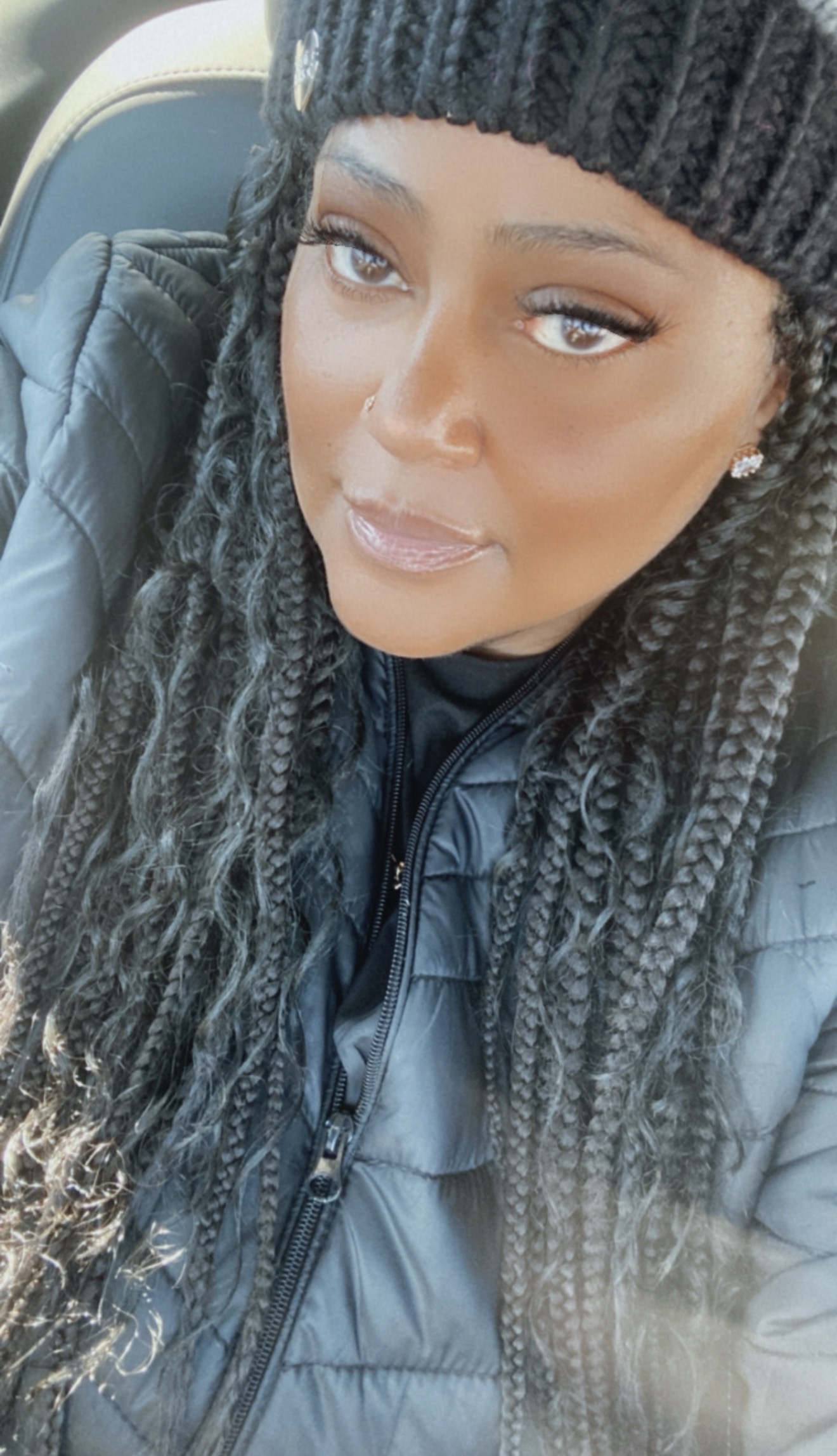 Woman with long braided hair wearing a black knit hat and a dark jacket, sitting in a vehicle.