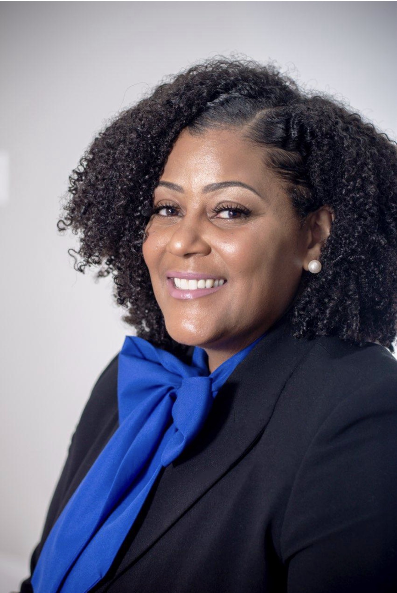 Portrait of a woman with curly hair, smiling, wearing a black blazer and a blue scarf, against a plain background.
