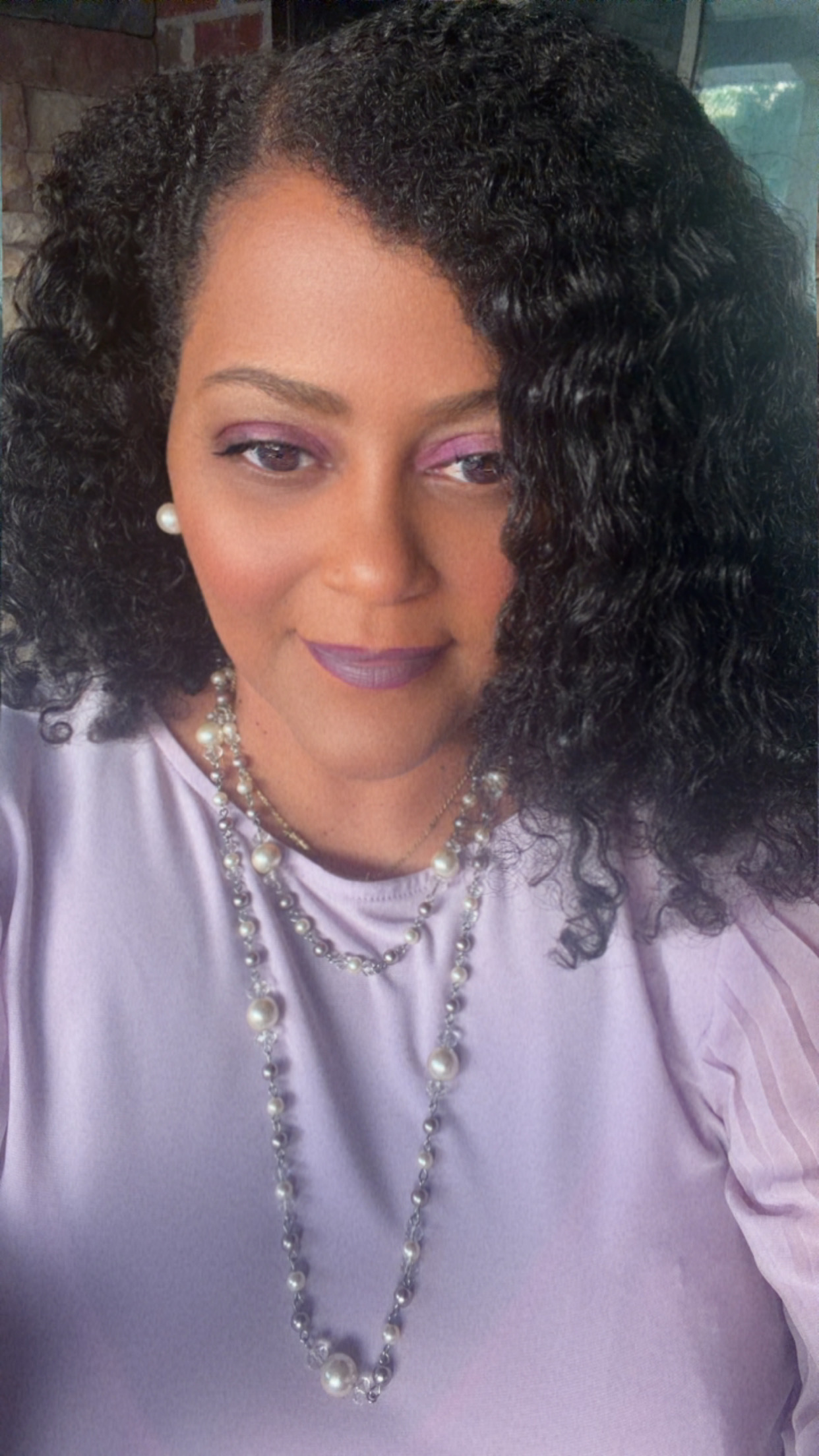 Woman with curly hair wearing pearl earrings and a pearl necklace, dressed in a light-colored top, looking at the camera.