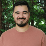 A smiling man with dark hair and a beard stands outdoors with green trees in the background.