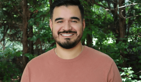 A smiling man with dark hair and a beard stands outdoors with green trees in the background.