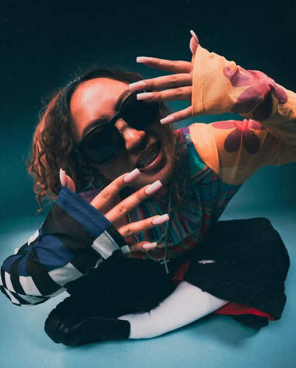 Person with curly hair wearing sunglasses, colorful hoodie, lying on the floor, making a hand gesture, dark background.