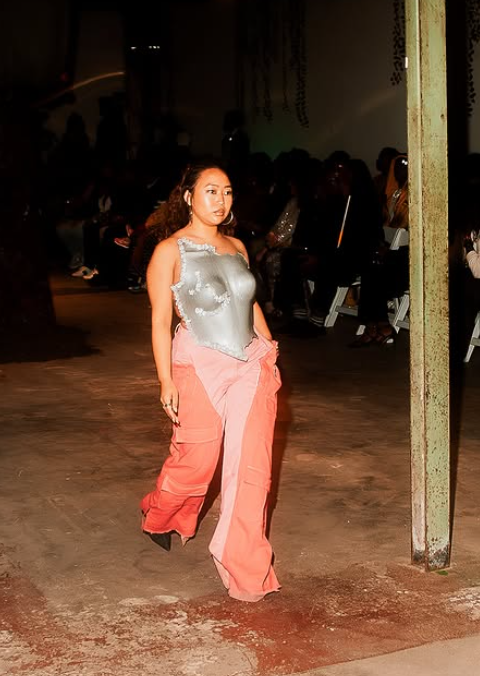 Woman walking on runway in silver top and pink wide-leg pants, audience seated in background, dim lighting.