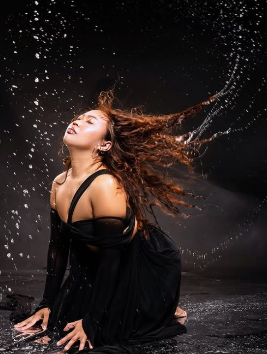 Woman with wet hair in a black dress kneeling on the ground, water splashing around her, dark background.