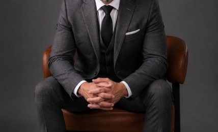 Man in a suit sitting on a chair with hands clasped, against a dark background.