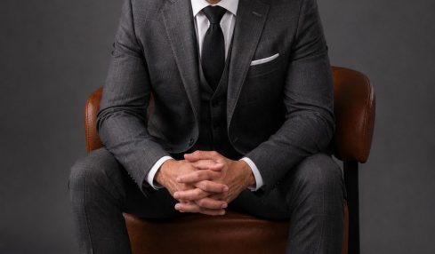 Man in a suit sitting on a chair with hands clasped, against a dark background.