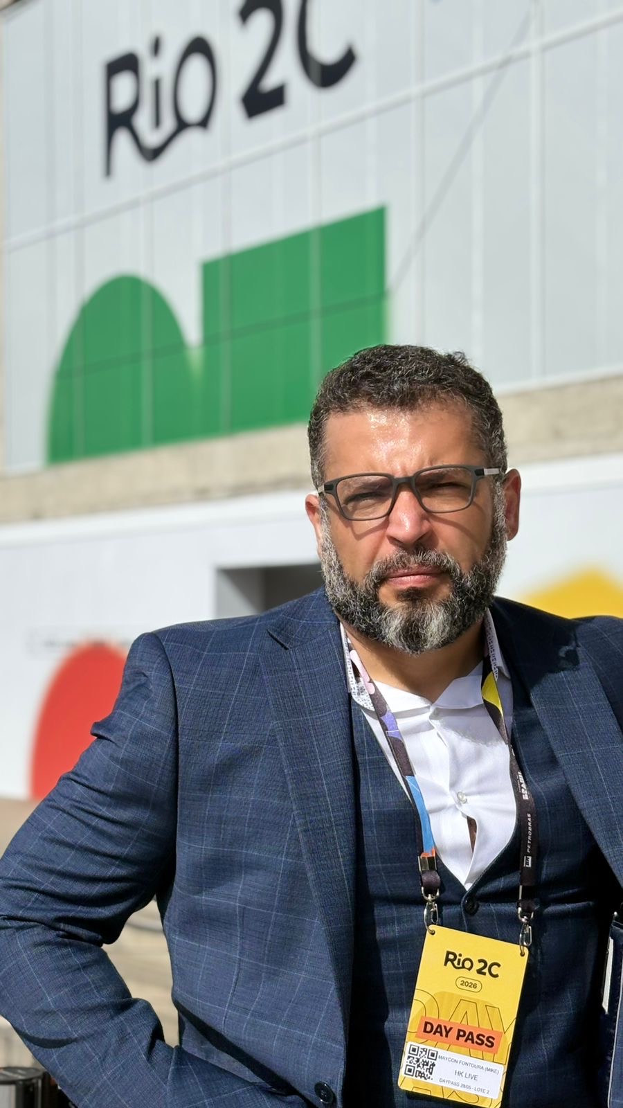 Man with glasses and beard wearing a suit and lanyard, standing outdoors in front of a Rio 2C sign.