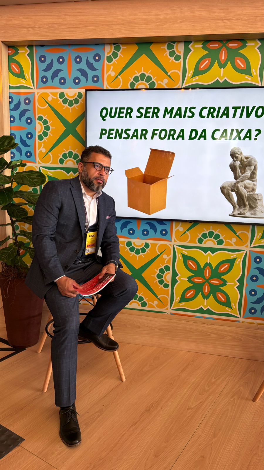 Man in suit sitting on a chair in front of a colorful tiled wall with a sign and a sculpture, holding a folder.