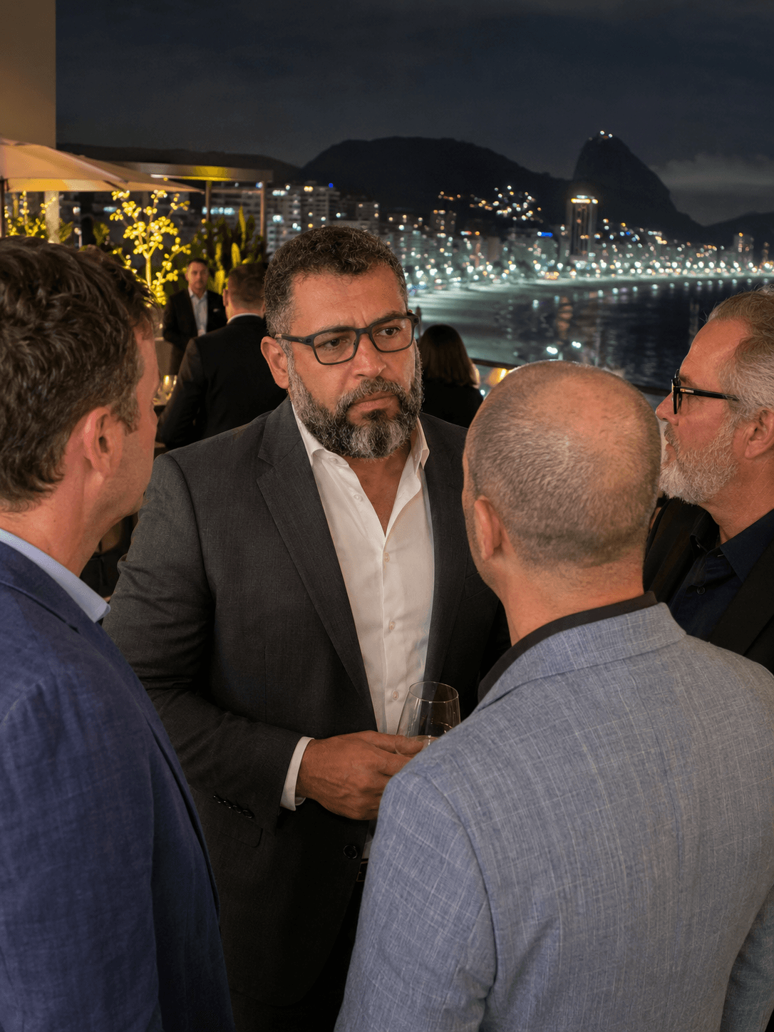 Group of four men in conversation at night with cityscape and water in background.