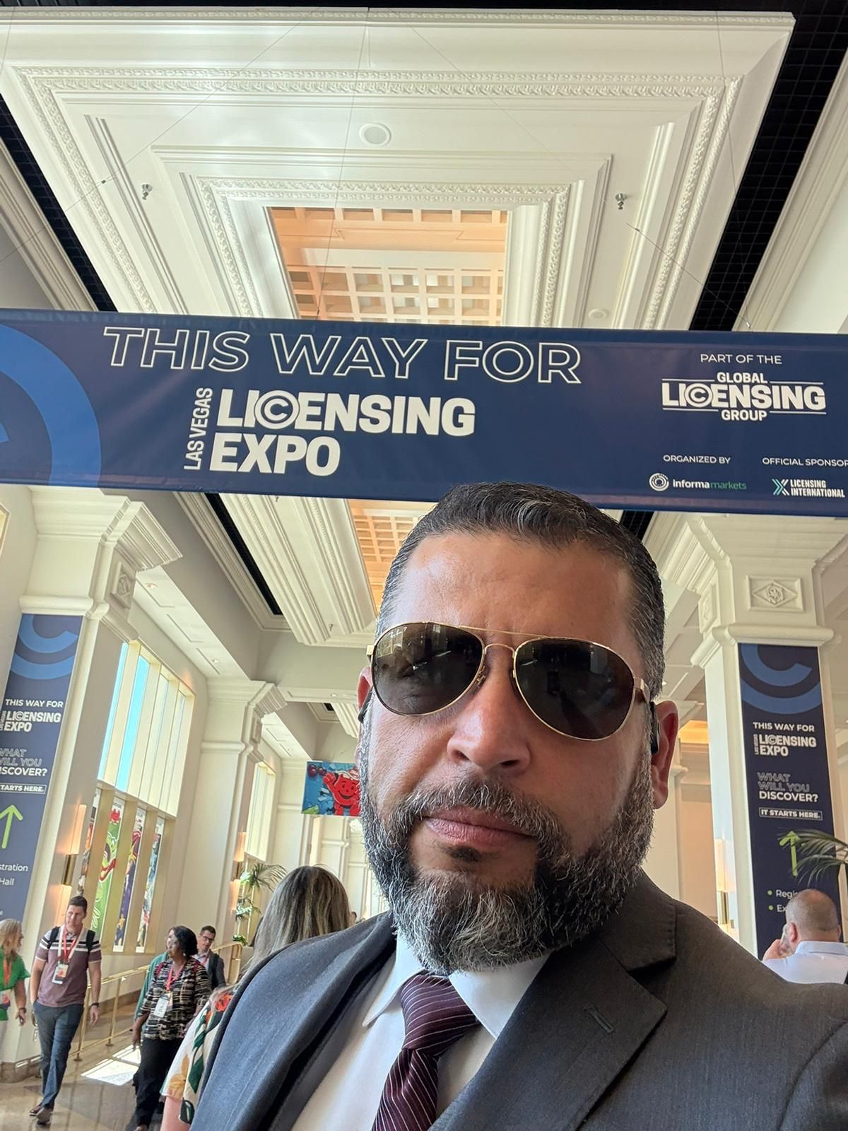 Man with sunglasses and beard in suit at licensing expo, indoor event with high ceiling and crowd.