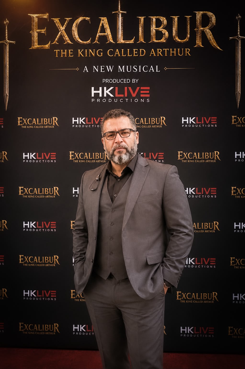 Man in gray suit and glasses standing in front of a black backdrop with gold text for 'Excalibur' musical, holding a phone.