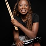 Smiling woman holding drumsticks and a drum, against a black background.