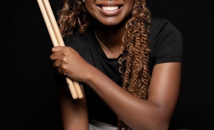 Smiling woman holding drumsticks and a drum, against a black background.