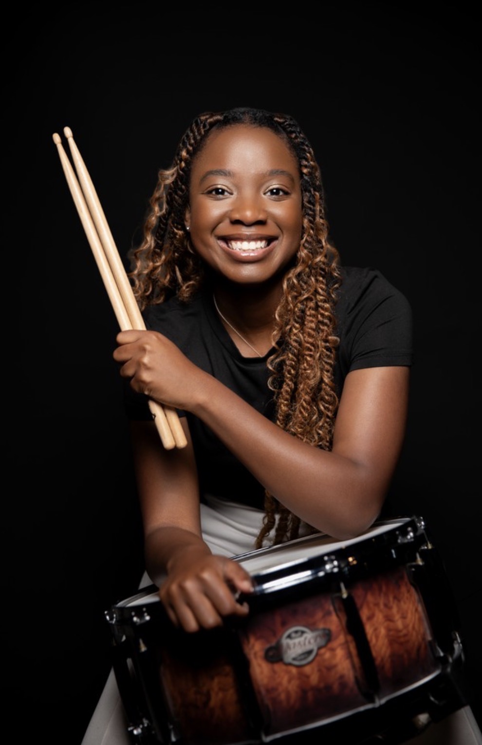 Smiling woman holding drumsticks and a drum, against a black background.
