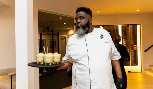 Chef in white uniform holding a tray with four glasses of dessert in a modern indoor setting.