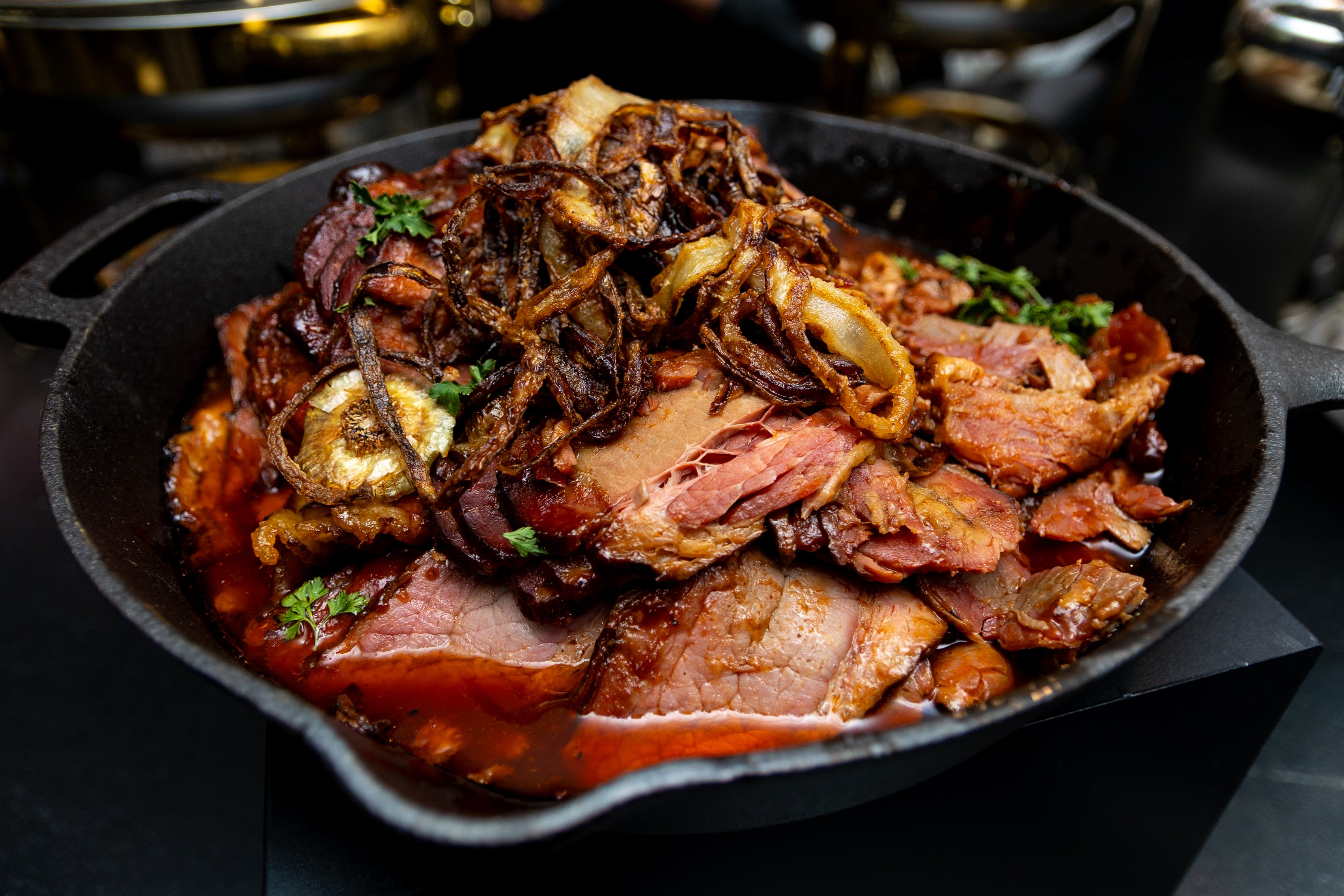 Cooked sliced meat with caramelized onions and herbs in a black skillet, garnished with green herbs.