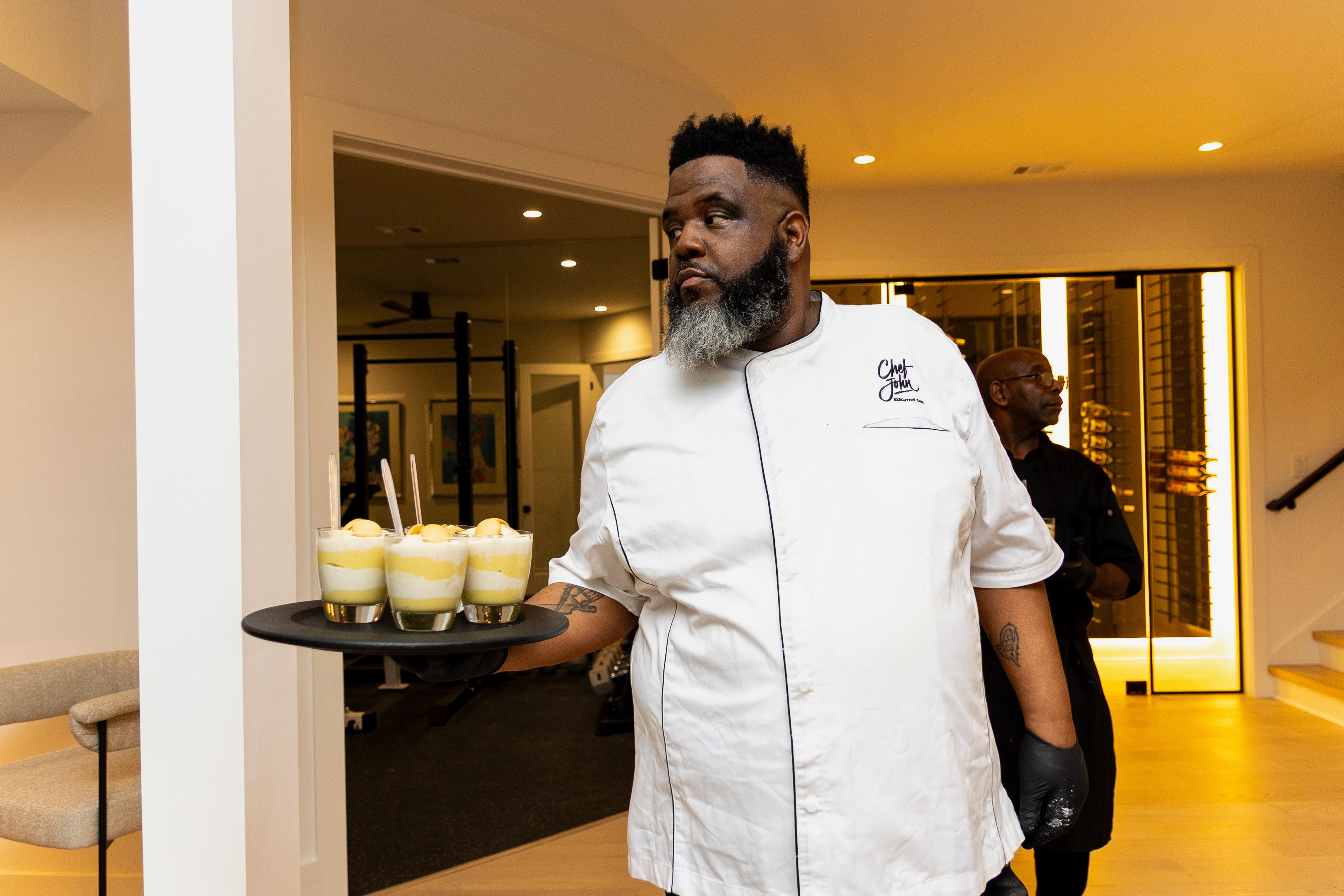 Chef in white uniform holding a tray with four glasses of dessert in a modern indoor setting.