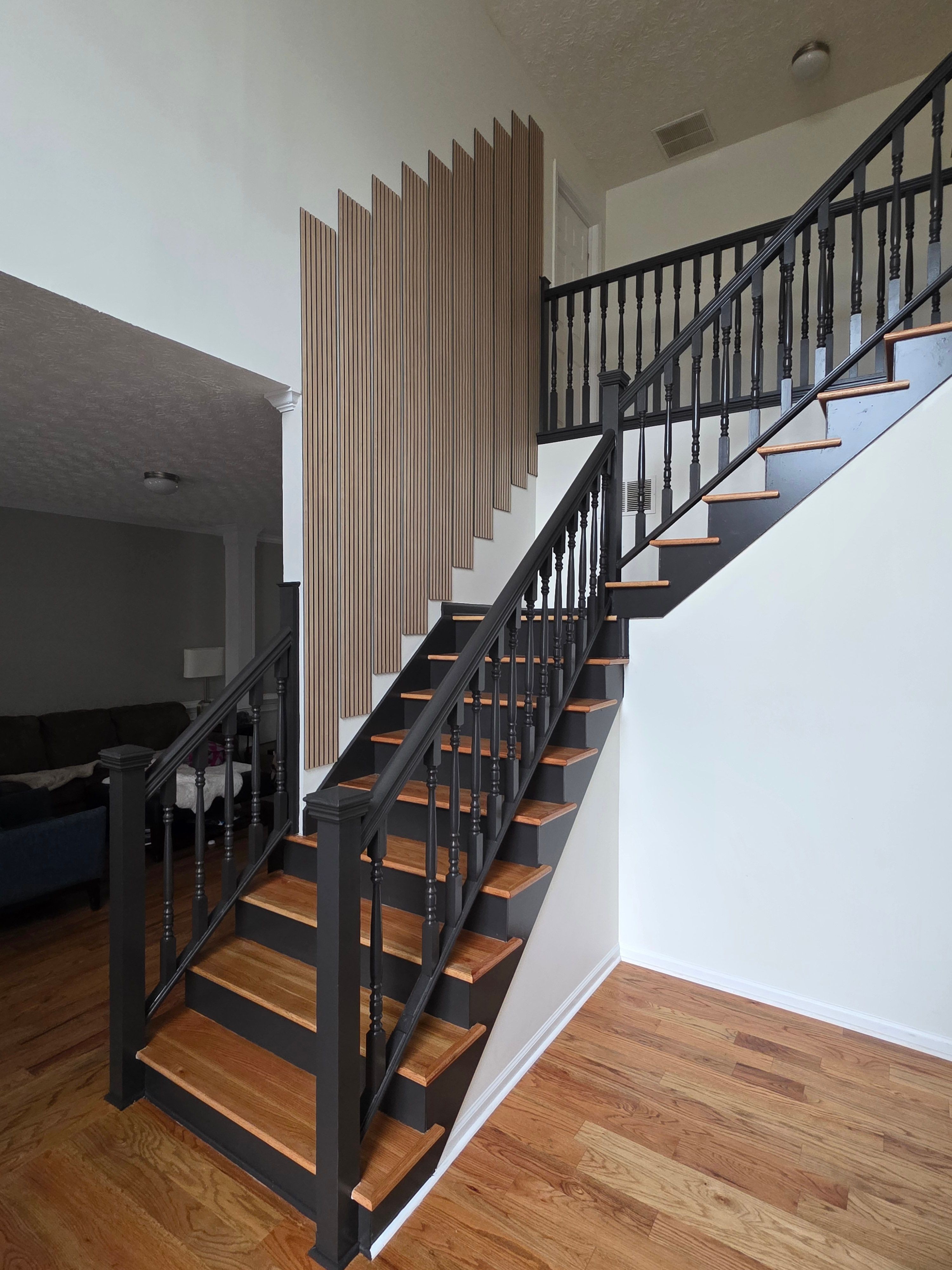 Staircase with black metal railing and wooden steps, viewed from below, with a white wall and wooden floor nearby.