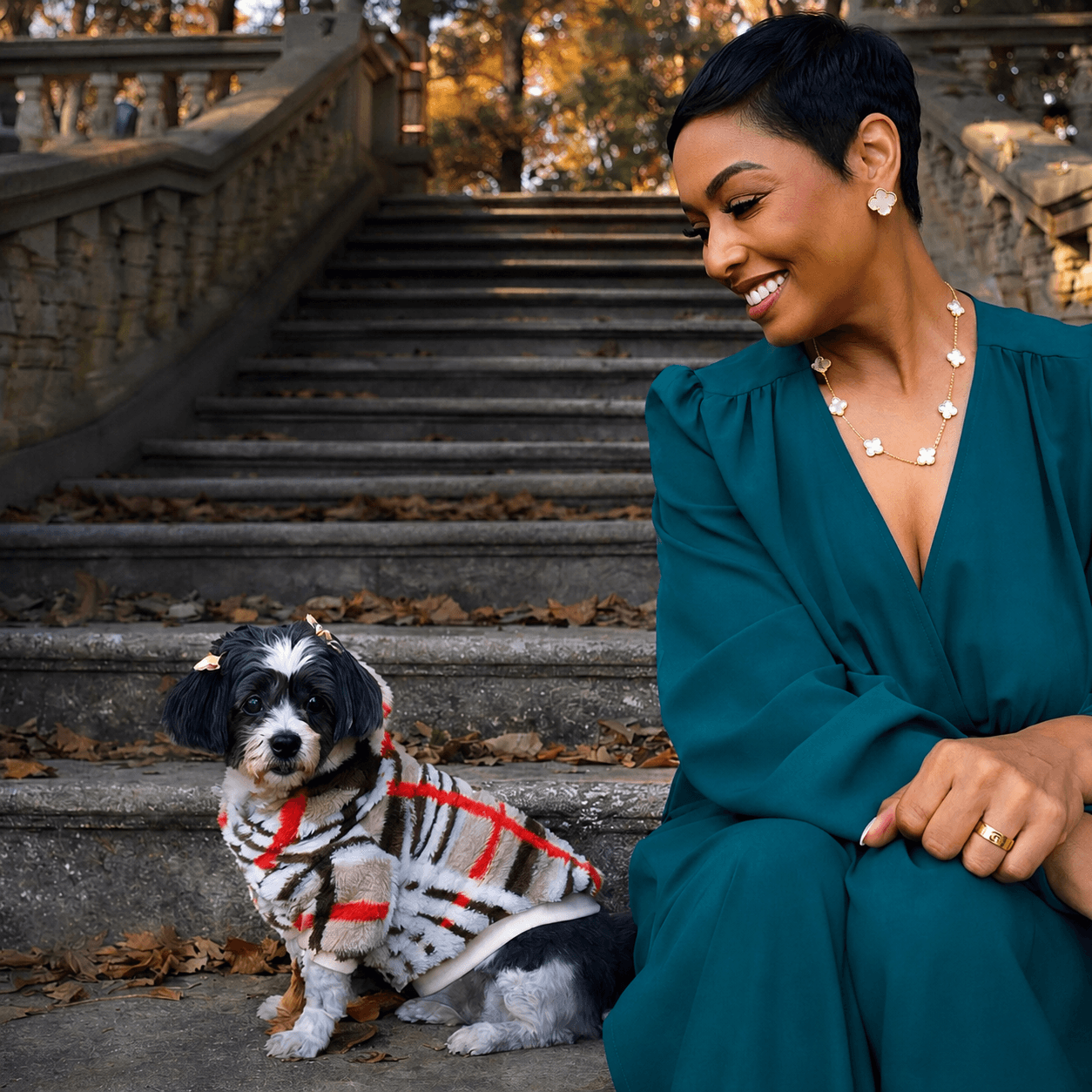 Woman with short black hair smiling at a small dog sitting on stairs outdoors, autumn leaves on the ground.
