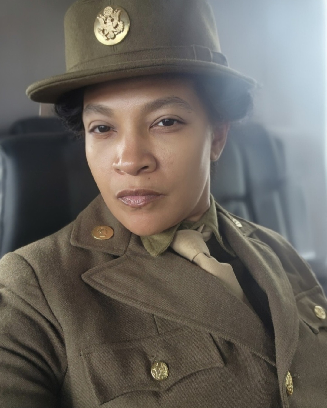 Person in military uniform with hat, looking directly at camera, seated inside vehicle, neutral expression.