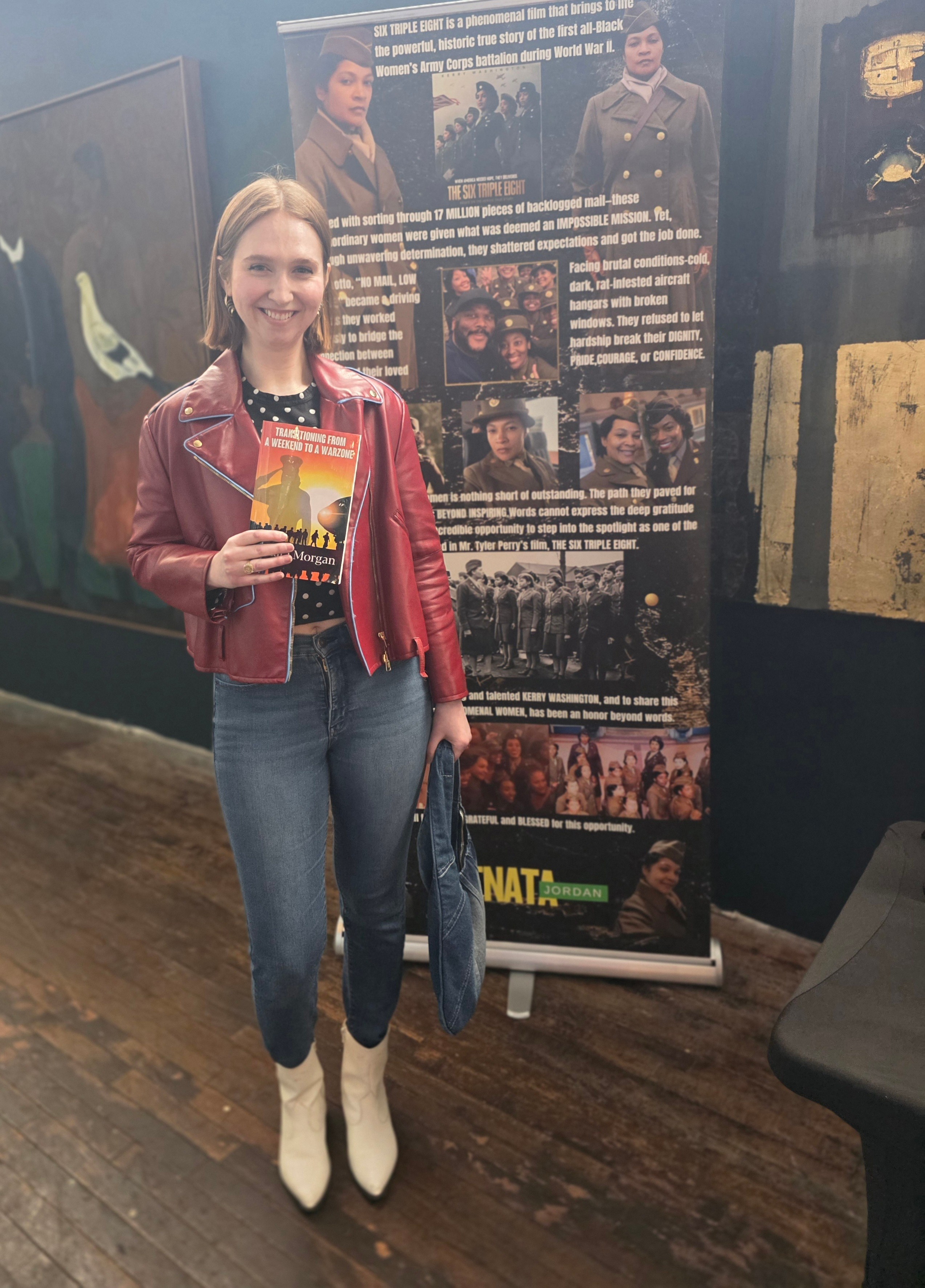 Woman with blonde hair in a red leather jacket and jeans holding a poster, standing indoors with display boards behind her.
