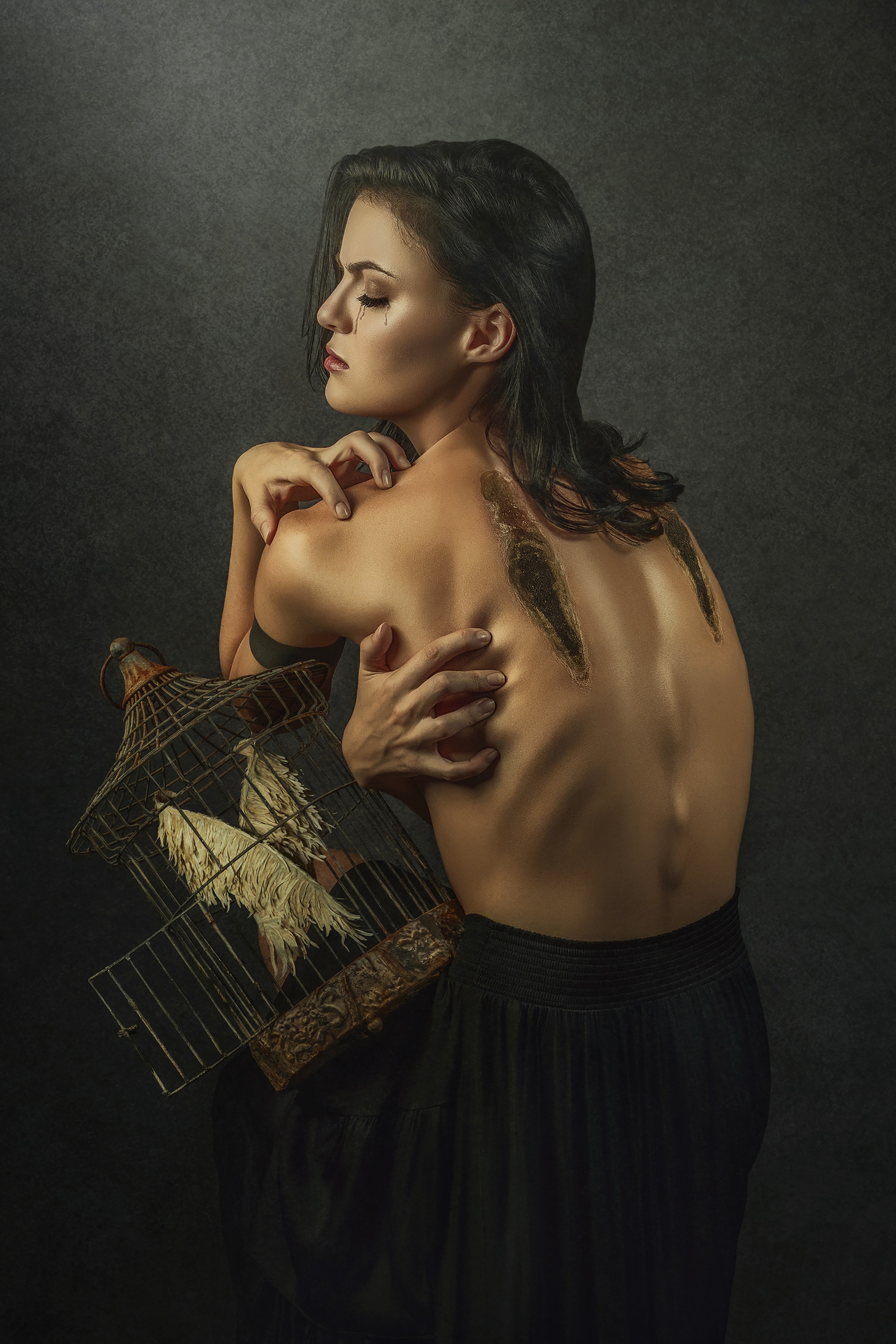A woman with dark hair and bare back holds a birdcage, looking down thoughtfully against a dark background.