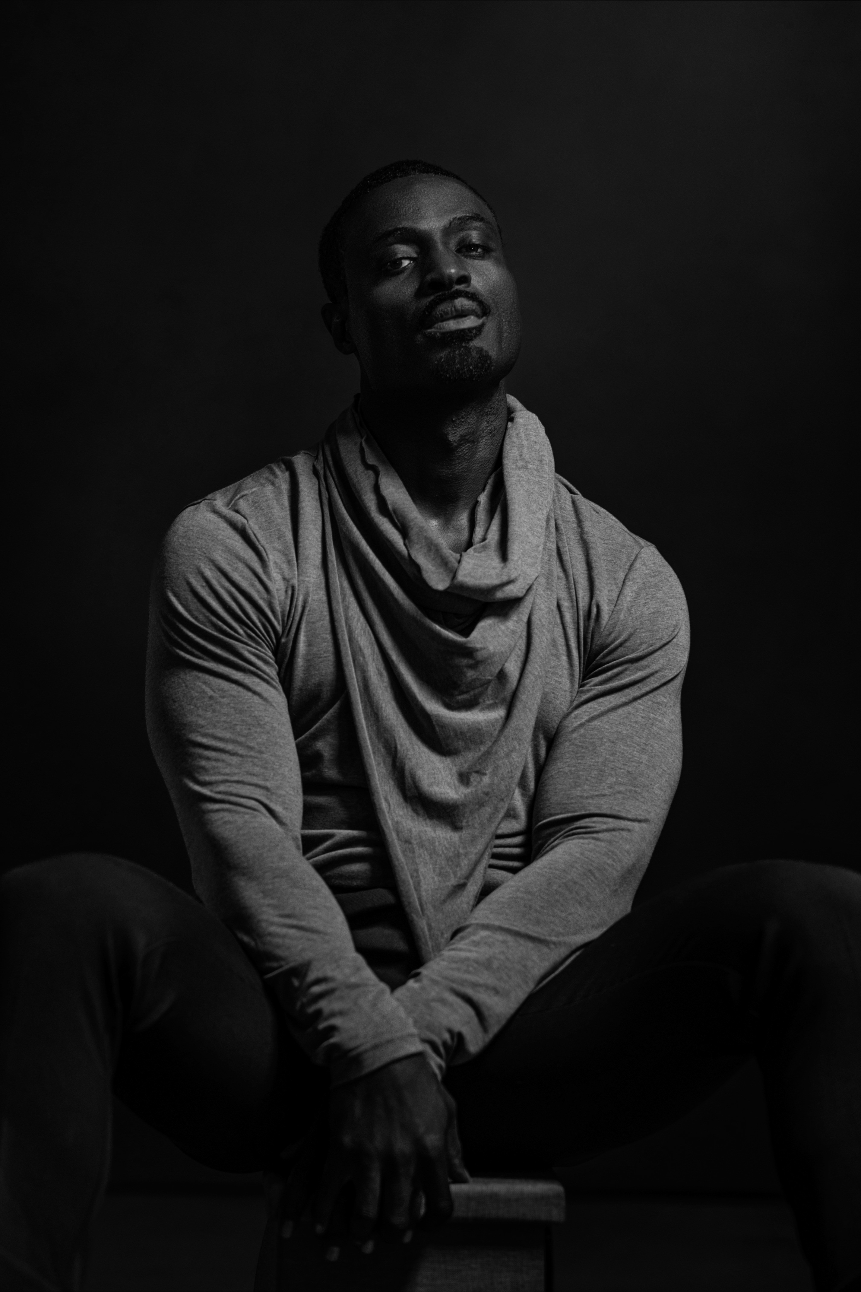 A man with a shaved head and beard sits with crossed legs, wearing a draped shirt against a dark background.