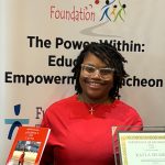 Smiling woman holding a book and a certificate in front of a banner about sickle cell disease foundation.