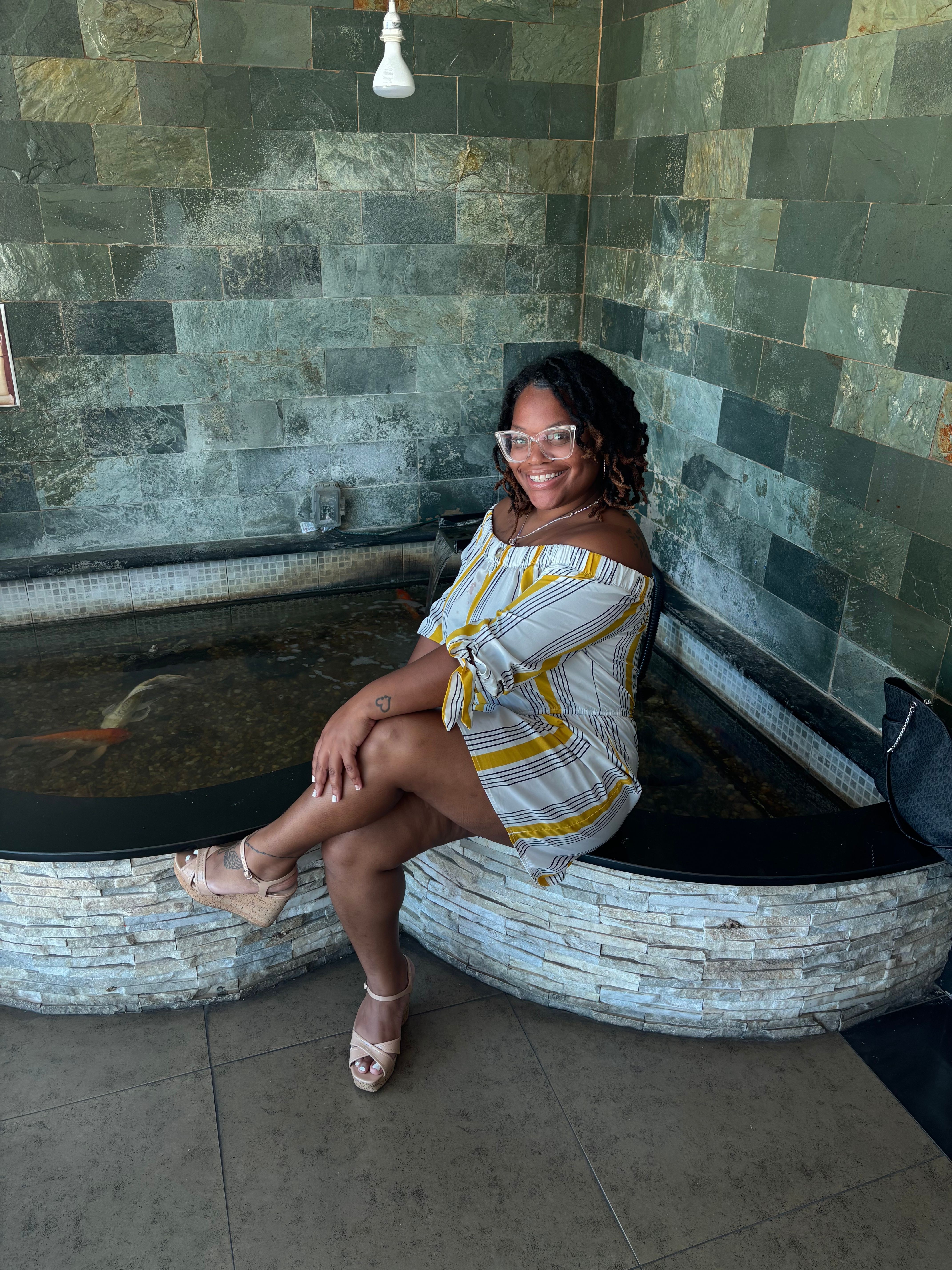 Woman sitting on a curved stone bench, wearing a yellow and white striped dress, glasses, and sandals, near a green tiled wall.