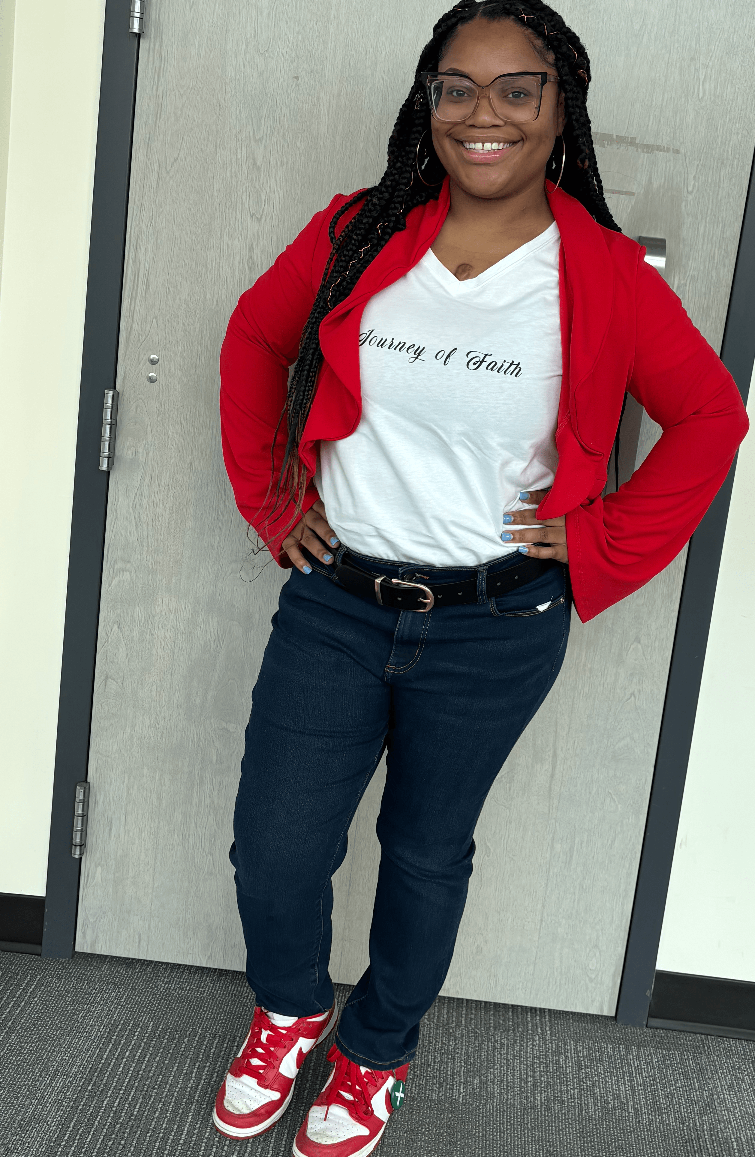 Smiling woman with glasses, wearing a white t-shirt, red jacket, dark jeans, and red sneakers, standing in front of a door.