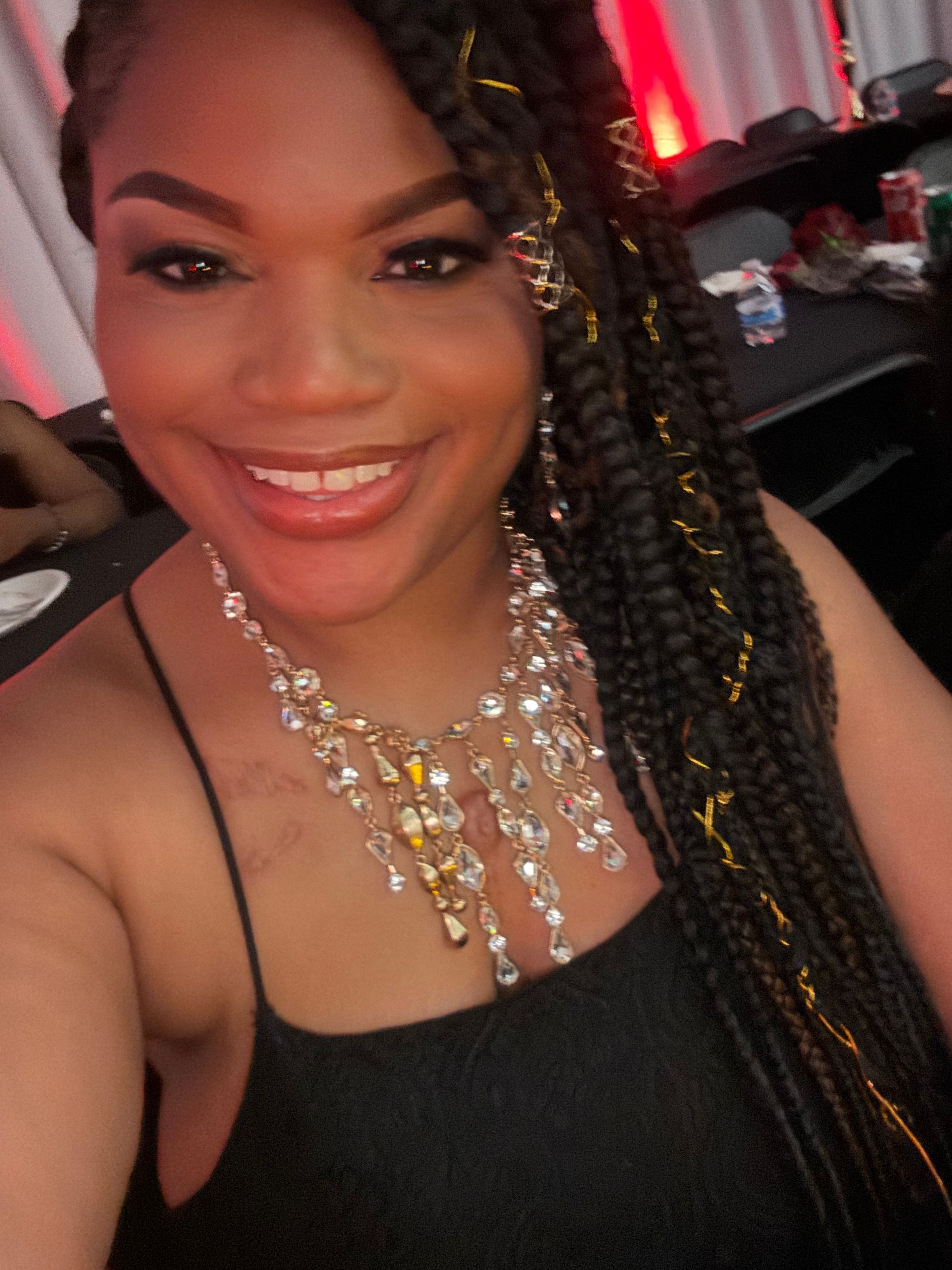 Smiling woman with braided hair wearing jewelry and a black top, sitting in a car.