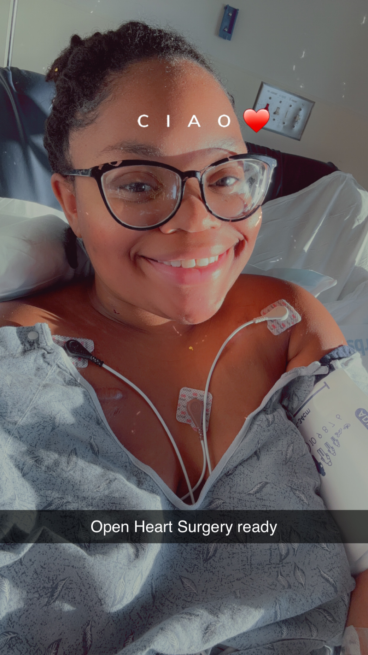 Smiling woman in hospital bed with medical tubes and glasses, text says 'CIAO' with a heart emoji, hospital room background.