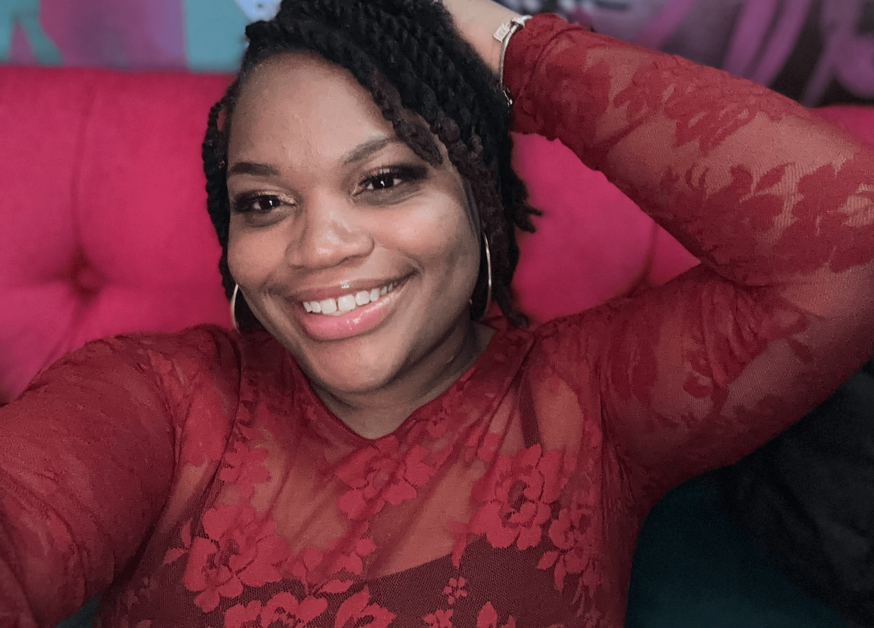 Smiling woman with braided hair in a red lace top, sitting on a pink couch.