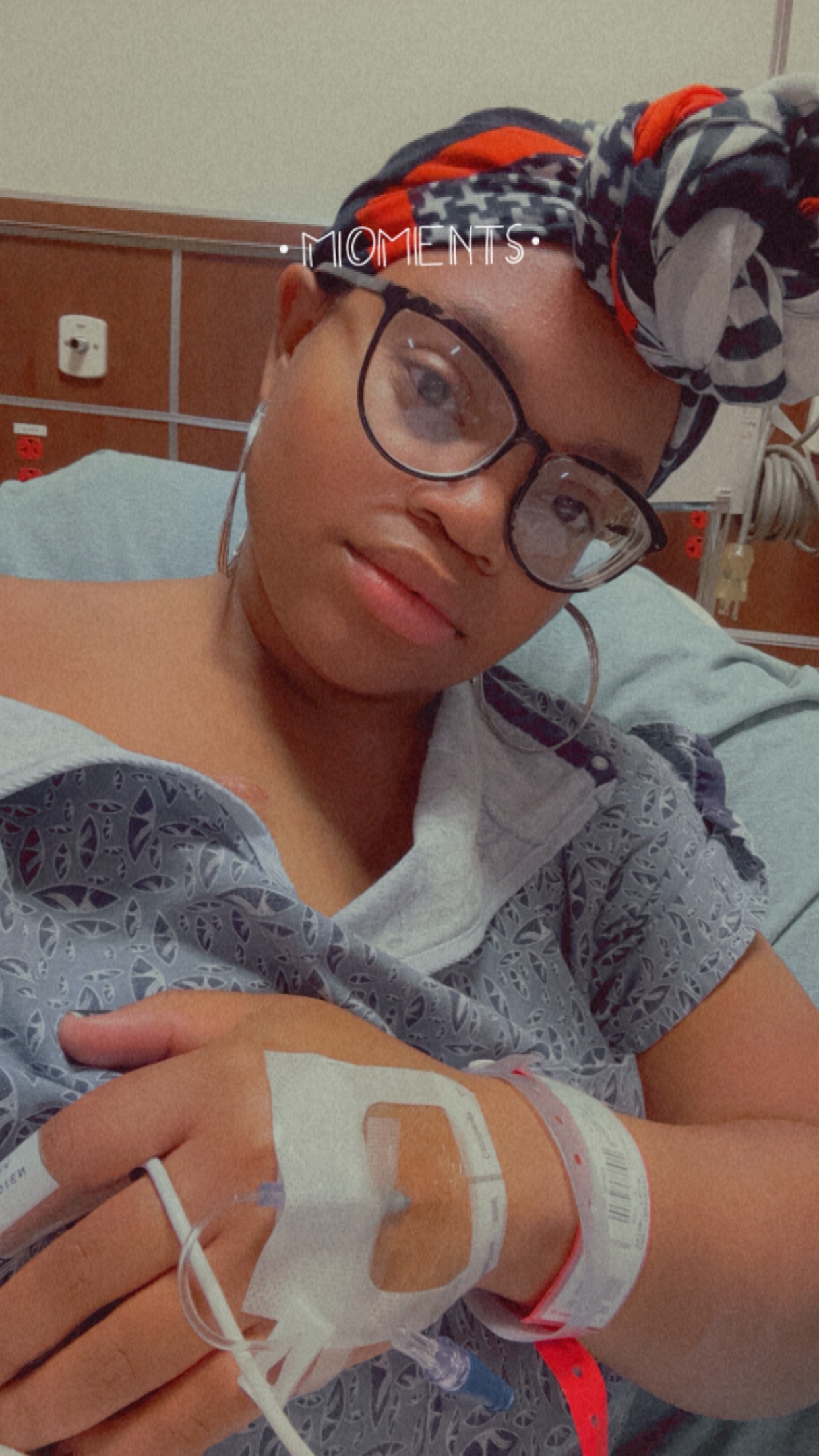 Young person in hospital bed with head bandage, wearing glasses, holding hand with medical device, looking at camera.