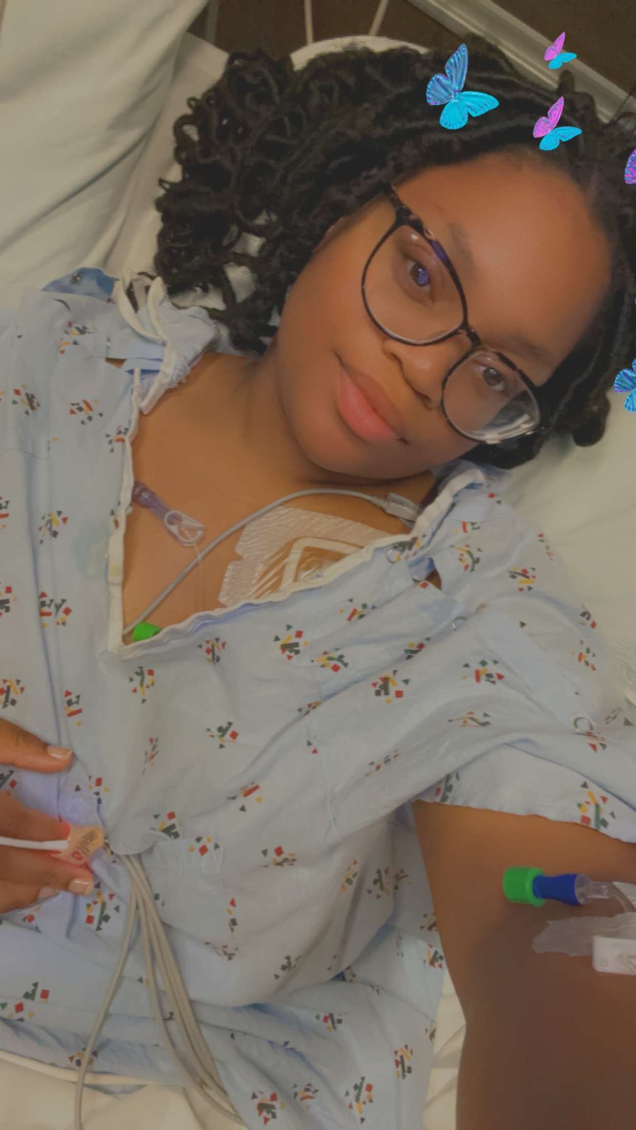 Young woman with glasses and curly hair lying down, wearing a floral top, with digital butterfly stickers above her head.