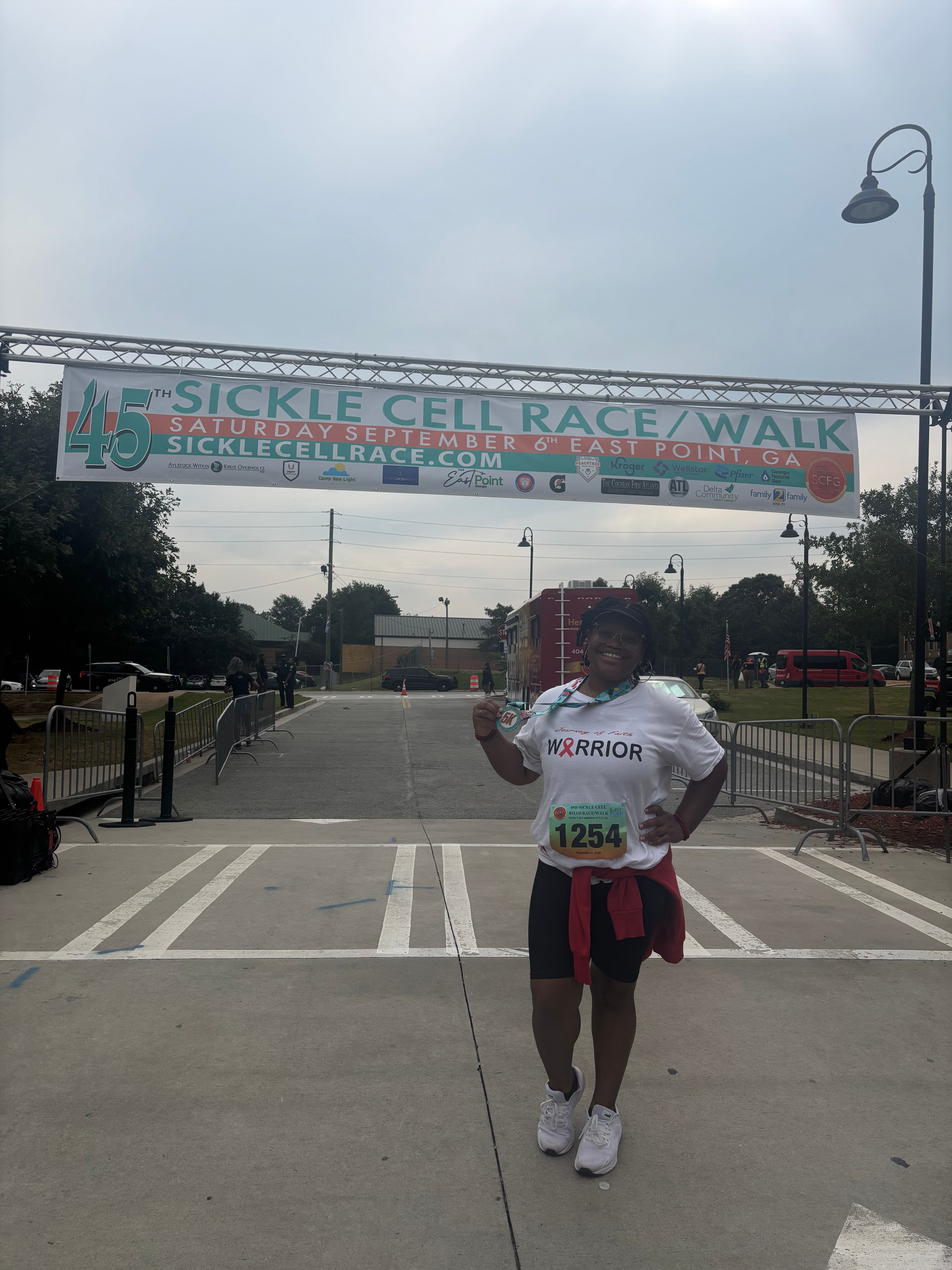 Person standing in a parking lot holding a sign, with a large banner in the background reading 'SICKLE CELL AWARENESS WALK'.